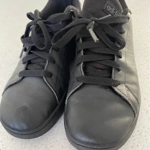 Svarta sneakers från Adidas - Klassiska helsvarta sneakers från Adidas med diskret perforering på sidorna och svart logga på häl och plös. Skorna har snörning, rund tå och platt sula med hexagonmönster undertill. Ovandelen är i skinnliknande material och insidan är vadderad för extra komfort.