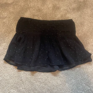 Svart kortkjol från Gina Tricot - Supersöt svart kortkjol från Gina Tricot med små prickiga detaljer och lätt volang. Kjolen har en bred, rynkad midja som sitter snyggt och ger en skön passform. Perfekt att matcha med en enkel topp eller hoodie.