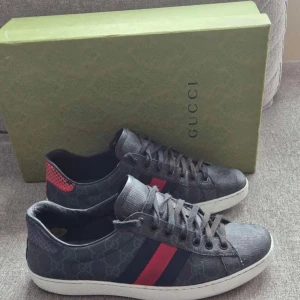 Gucci sneakers med röd/blå rand - Snygga svarta Gucci sneakers med klassiskt GG-mönster, röd och marinblå rand på sidan och vit sula. Skorna har snörning och detaljer i skinn, samt en röd detalj i ormskinnslook på hälen. Perfekta för dig som vill sticka ut med lyxig streetstyle.