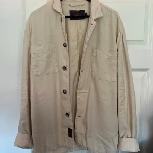 Beige overshirt från Morris, storlek M - Stilren beige overshirt från Morris med klassisk krage, två stora bröstfickor och knäppning framtill. Skjortan är i bomull och linne och har långa ärmar med manschett. Perfekt att bära öppen över en t-shirt för en avslappnad look. Knappt använd.