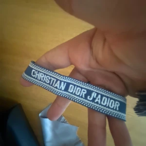 Christia diorr armband - Snyggt blått armband från Christian Dior med texten 'J'ADIOR' broderad i vitt. Armbandet har en cool, casual look och är gjort i tyg med vita detaljer längs kanterna. Perfekt accessoar för att lyfta din outfit.