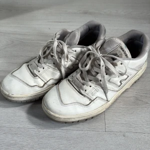 Vita New Balance 550 sneakers - Säljer ett par klassiska New Balance 550 sneakers i vitt med grå detaljer. Skorna har en chunky siluett, snörning och platt sula. Ovandelen är i skinn och mesh för en sportig vibe. Perfekta för dig som gillar retrostil och streetwear.