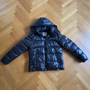 Svart Moncler maya - Snygg svart pufferjacka från Moncler med glansig finish och klassisk logga på ärmen. Jackan har huva, dragkedja framtill och två fickor med dragkedja. 