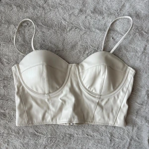 Beige korsett-topp med smala axelband - Snygg beige korsett-topp i syntetmaterial med smala justerbara axelband och markerade kupor. Toppen har en croppad passform och dragkedja bak för enkel påklädning. Perfekt för att lyfta vilken outfit som helst.