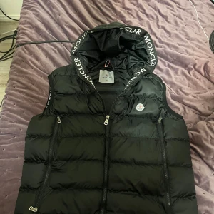 Moncler vest  - Säljer en svart dunväst från Moncler med stor huva och tydlig logga på bröstet. Västen har två dragkedjefickor framtill och dragkedja i mitten. Moncler-tryck längs huvkanten ger en clean och sportig vibe. Perfekt för lager-på-lager-stil.