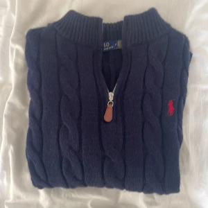 Mörkblå kabelstickad tröja - Kabelstickad mörkblå tröja  med halv dragkedja och ribbad krage. Klassisk röd logga broderad på bröstet. Tröjan är långärmad och gjord i mjuk ull, perfekt för kyliga dagar.