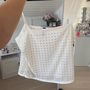 Vit glittrig linne-topp från H&M Divided - Supersnygg vit linne-topp från H&M Divided med tunna axelband och massor av silvriga glittriga nitar framtill. Perfekt för dig som vill sticka ut lite extra! Toppens material känns mjukt och lätt, och den har en enkel, rak passform.