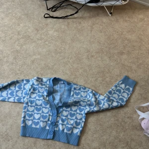 Blå kofta med hjärtmönster och knappar - Supergullig blå kofta med vita hjärtan över hela tröjan. Croppad modell med bred ribbad mudd och tre stora knappar framtill. Perfekt för dig som vill ha en söt och trendig look. Mjuk och stickad känsla, långärmad och v-ringad.