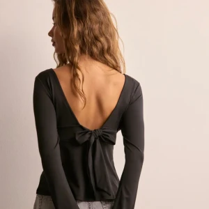 Svart långärmad topp med rosett Nelly - Svart långärmad topp från Nelly med djup v-ringning fram och öppen rygg med en snygg knytrosett. Toppens material är mjukt och stretchigt, vilket gör den bekväm och smickrande. Perfekt för dig som gillar stilrena detaljer och en cool rygg.