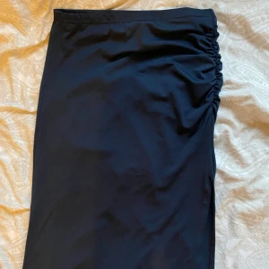 Svart långkjol med slits och rynk - Svart långkjol med snygg rynkdetalj på ena sidan och hög slits för extra edge. Kjolen har en enkel, stilren design och är gjord i ett mjukt, stretchigt material som sitter skönt. Perfekt för dig som gillar minimalistisk stil med en twist.