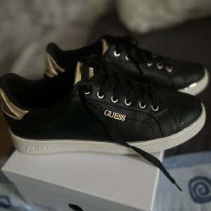 Svarta sneakers från Guess - Snygga svarta sneakers från Guess med gulddetaljer och präglat mönster. Skorna har snörning och en vit sula som ger en stilren look. Perfekta för att lyfta din outfit. Använda en gång bra och bekväma skor säljer pga fel med storlek 