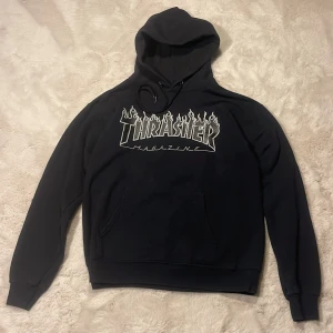 Svart Thrasher hoodie  - Svart hoodie från Thrasher med ikoniskt flames-tryck på bröstet. Klassisk känguruficka, huva med snörning och ribbade muddar. Materialmix av bomull och polyester gör den både mjuk och skön. Perfekt för dig som gillar streetwear och skatekänsla.