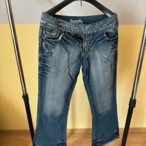 låg midjade jeans från Bench - Snygga blå jeans från Bench som är lågmidjade och har lätt slitna detaljer. Klassisk femficksmodell med Bench-logga på baksidan. Jeansen har en flared passform och är tillverkade i denim med coola slitningar framtill och baktill. Aldrig använda då dom var för små för mig