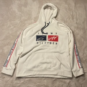 Vit hoodie från Tommy Hilfiger - Snygg vit hoodie från Tommy Hilfiger med stor logga framtill i blått och rött. Svarta snören i huvan och breda band med Tommy Hilfiger-tryck längs ärmarna. Klassisk känguruficka och mjukt material.