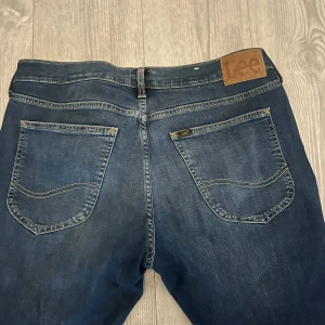 Lee Luke jeans W32 L32 mörkblå - Snygga mörkblå Lee Luke jeans med klassisk femficksdesign och gul kontrastsöm. Jeansen har en slim passform och är tillverkade i mjukt denimtyg. Perfekta för dig som gillar en modern och stilren look.