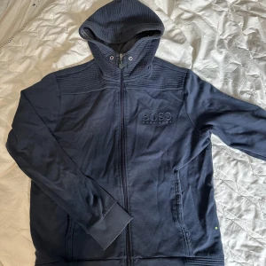 Mörkblå hoodiejacka från Hugo Boss - Mörkblå hoodiejacka från Hugo Boss med broderad logga på bröstet. Jackan har dragkedja, huva med snäppknapp och ribbade detaljer vid axlar, ärmslut och nederkant. Tillverkad i mjuk bomull för en skön och avslappnad stil.  Dock använt skick,  kolla bilder för skick 