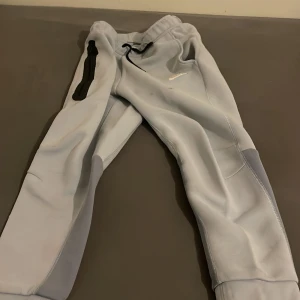 Ljusgrå mjukisbyxor från Nike - Nike mjukisbyxor i ljusgrått med svarta detaljer och dragkedjeficka på ena benet. Byxorna har elastisk midja med snörning och förstärkta partier vid knäna. Perfekta för chill eller träning, riktigt sköna och stilrena.