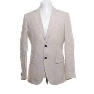 Beige linnékavaj från Bläck - Stilren beige kavaj från Bläck i 100% linne. Klassisk passform med två knappar framtill, fickor med lock och slits bak. Perfekt för en fräsch och avslappnad look under varmare dagar. Skandinavisk design med snyggt fall och lätt struktur. Nypris 1999kr knappt använd 