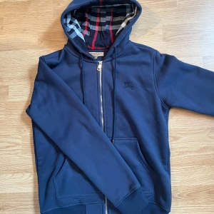 Mörkblå Burberry hoodie med dragkedja - Snygg marinblå hoodie från Burberry med klassiskt rutigt foder i huvan. Tröjan har dragkedja framtill, snörning i huvan och broderad logga på bröstet. Tillverkad i mjuk bomull och har praktiska fickor framtill. Perfekt för dig som gillar stilrena och exklusiva plagg.