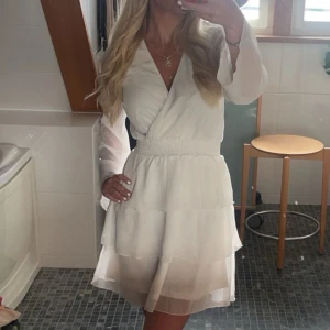 Vit omlottklänning med volang - Superfin vit kortklänning med omlottliv och v-ringning. Klänningen har långa, transparenta ärmar och volangdetaljer i kjolen. Resår i midjan ger en snygg passform. Perfekt för sommarens alla tillfällen!