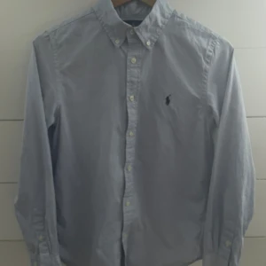 Blå skjorta från Ralph Lauren - Klassisk ljusblå skjorta från Ralph Lauren med button-down krage och broderad logga på bröstet. Skjortan har långa ärmar och knäppning framtill. Perfekt för dig som gillar stilrena och tidlösa plagg.