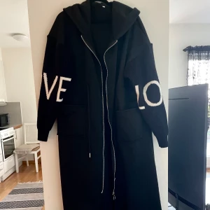 Svart lång hoodie-kappa med tryck - Lång svart kappa / tröja med huva och dragkedja framtill. Ärmarna har stora vita bokstäver som bildar ordet 'LOVE'. Två stora fickor framtill och avslappnad passform. Perfekt för dig som gillar streetstyle och gillar bekvämt.