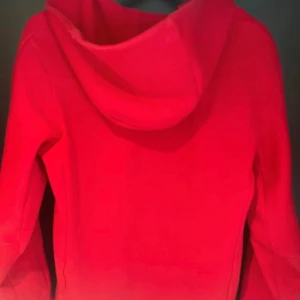Röd Nike vindjacka med huva XS - Nike vindjacka i rött med svarta detaljer och dragkedja framtill. Jackan har huva, lång ärm med ficka och dragkedja på ärmen samt svarta ränder över bröstet. Perfekt för dig som gillar sportig stil och vill sticka ut.