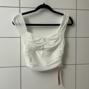 Vit croppad topp från SHEIN  - Supersnygg vit croppad topp från SHEIN med breda axelband och rynkade detaljer framtill och baktill. Toppens material är mjukt och lätt, perfekt för att skapa en trendig look. Passar perfekt till både jeans och kjol för en cool stil.