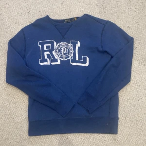 Blå sweatshirt från Polo Ralph Lauren - Mörkblå sweatshirt från Polo Ralph Lauren med stor vit RL-logga framtill. Klassisk crewneck-modell med ribbade muddar vid ärmslut och nederkant. Skön bomullskvalitet och långärmad passform, perfekt för en chill och stilren look. Perfekt inför sena sommar kvällar när de blir lite kyligare 