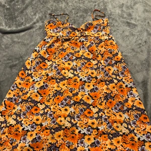 Blommig sommarklänning med smala axelband - Superfin kort klänning med smala axelband och färgglatt blommönster i orange, gult, lila och svart. Perfekt för varma dagar och har en lätt, flowy passform. Snygg v-ringning och skön bomullskänsla.