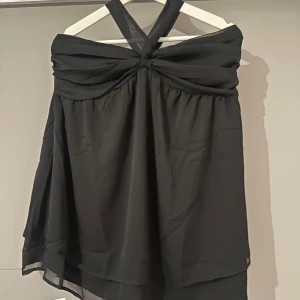 Svart kort klänning från Boohoo - Supersnygg svart kort klänning från Boohoo med omlott-detalj upptill och breda axelband som korsas i nacken. Klänningen har ett lätt, flowy tyg och dragkedja bak för enkel påklädning. Perfekt för dig som vill sticka ut med en stilren look.