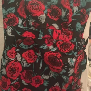 Svart klänning med röda rosor - Supersnygg svart klänning med stora röda rosor och gröna blad. Klänningen har ett blommigt mönster och ett knytband i midjan som ger en fin siluett. Perfekt för dig som vill sticka ut med färg och mönster.