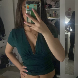 Mörkgrön v-ringad topp - Snygg mörkgrön topp med djup v-ringning och korta ärmar. Modellen är figurnära och croppad, vilket ger en modern och trendig look. Perfekt för dig som vill sticka ut med en stilren men ändå vågad topp. Pris kan diskuteras