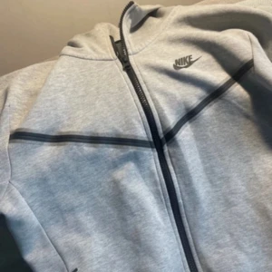 Grå nike tech från Nike  - Snygg grå zipjacka från Nike i storlek XS. Jackan har hel dragkedja framtill, svarta detaljer och diskret Nike-logga på bröstet. Tillverkad i mjukt material som passar perfekt till chill eller träning. Enkel och stilren design med sportig vibe.