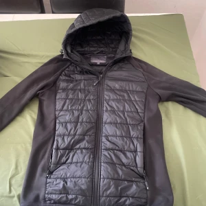 Svart pufferjacka från Cross Sportswear - Svart pufferjacka från Cross Sportswear med huva och hel dragkedja. Perfekt för kyliga dagar och enkel att matcha med det mesta. Pris går o diskutera använd ett fåtal gånger. 
