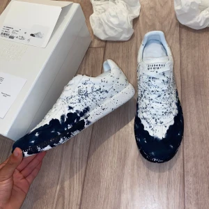 Maison Margiela paint  - Unika sneakers från Maison Margiela i vitt skinn med svart färgstänk och coolt splattermönster. Klassisk låg modell med snörning och rund tå. Kommer med originalbox och dustbags. Perfekt för dig som vill sticka ut med en konstnärlig vibe 