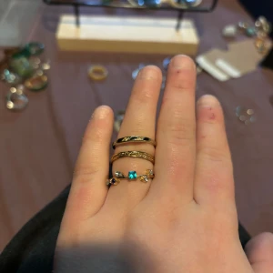 Set med tre guldiga ringar med stenar - Säljer ett set med tre snygga guldiga ringar. En ring har ett tvinnat mönster, en har glittrande små stenar runt hela och den tredje är dekorerad med små vita och blå stenar samt en fyrkantig blå sten i mitten. Perfekt för att mixa och matcha på fingrarna.