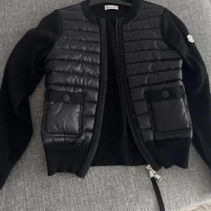 Svart cardigan jacka från Moncler - Snygg svart cardigan jacka från Moncler med quiltad framsida och ärmar i stickad ull. Jackan har två fickor, qr kod, nfc och taggar!