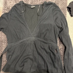 Svart omlottblus från Gina Tricot - Svart omlottblus från Gina Tricot med v-ringning och långa ärmar. Blusen har knytband i midjan och är tillverkad i ett lätt, strukturerat tyg som ger en snygg och avslappnad look. Perfekt att styla med jeans eller kjol.