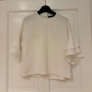 Vit volangblus från Zara Woman - Supersnygg vit blus från Zara Woman med rund hals och korta ärmar med dubbla volanger. Blusen har en rak passform och är tillverkad i ett lätt, flowy material som ger en elegant vibe. Perfekt att styla med jeans eller kjol för en clean look.