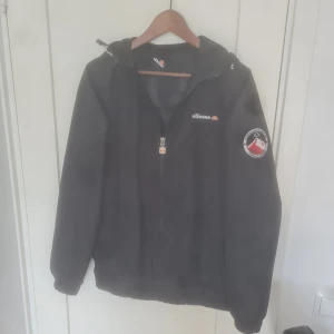 Svart vindjacka från Ellesse med patch - Svart vindjacka från Ellesse med huva och dragkedja framtill i mycket bra skick. Jackan har Ellesse-logga på bröstet och en snygg broderad patch på ärmen. Perfekt för dig som gillar sportig och clean stil. Storlek M. Bara att skriva vid frågor