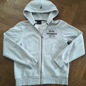 Ljusgrå hoodie Peak Performance XL - Ljusgrå hoodie från Peak Performance i storlek XL. Klassisk modell med dragkedja, huva och två fickor framtill. Broderad logga på bröstet och liten detalj på huvan. Mjuk bomullsblandning, perfekt för chill dagar.