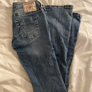 True Religion jeans  - Snygga blå jeans från True Religion med klassisk kontrastsöm och ikonisk bakficka. Modellen är utsvängda nere vid anklarna, silverfärgade knappar, baby rosa i sömmen och dragkedja. Perfekta för dig som gillar streetstyle och vill sticka ut med ett coolt märke.