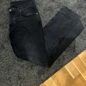 Gråa nudie jeans - Säljer ett par gråa jeans med breda ben och klassisk femficksdesign. Jeansen har en mörk tvätt och raka, vida ben som ger en avslappnad och trendig look. Perfekta till sneakers eller boots. 32/30