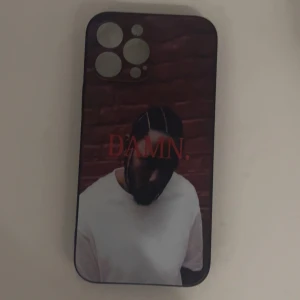 Kendrick Lamar Mobilskal - Passar till IPhone 13 Pro Max! Bilderna visar ett mobilskal med motivet från Kendrick Lamars album 'DAMN.' och en bild på artisten..