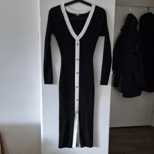 Svart lång stickad klänning med knappar - Snygg svart långklänning med vit kant längs v-ringningen och hela knappslån. Klänningen är ribbstickad, har långa ärmar och guldiga knappar framtill. Den är figurnära och har en liten slits nertill för extra rörelsefrihet.