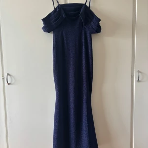 Mörkblå glittrig långklänning - Snygg mörkblå långklänning med glittrigt tyg och smala axelband. Klänningen har en unik volangdetalj över axlarna som ger en cool och elegant look. Perfekt för dig som vill sticka ut med en stilren och trendig outfit.