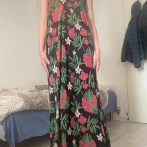 Blommig långklänning med slits - Snygg långklänning med tropiskt blommönster i rött, grönt och vitt på svart botten. Klänningen har smala axelband, smock upptill och en hög slits på ena sidan. Perfekt för sommarens varma dagar och ger en chill vibe.