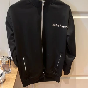  Palm Angels hoodie  - Snygg svart track jacket från Palm Angels med vita detaljer och dragkedja. Jackan har långa ärmar med vita ränder längs sidorna och en hög krage. Perfekt för en sportig look.