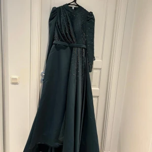 Mörkgrön långklänning med pärlor - Elegant mörkgrön långklänning med glittrande pärldetaljer över hela överdelen och ärmarna. Klänningen har lång ärm, markerad midja med bälte och ett vackert fall. Perfekt för dig som vill sticka ut med en lyxig och sofistikerad look.